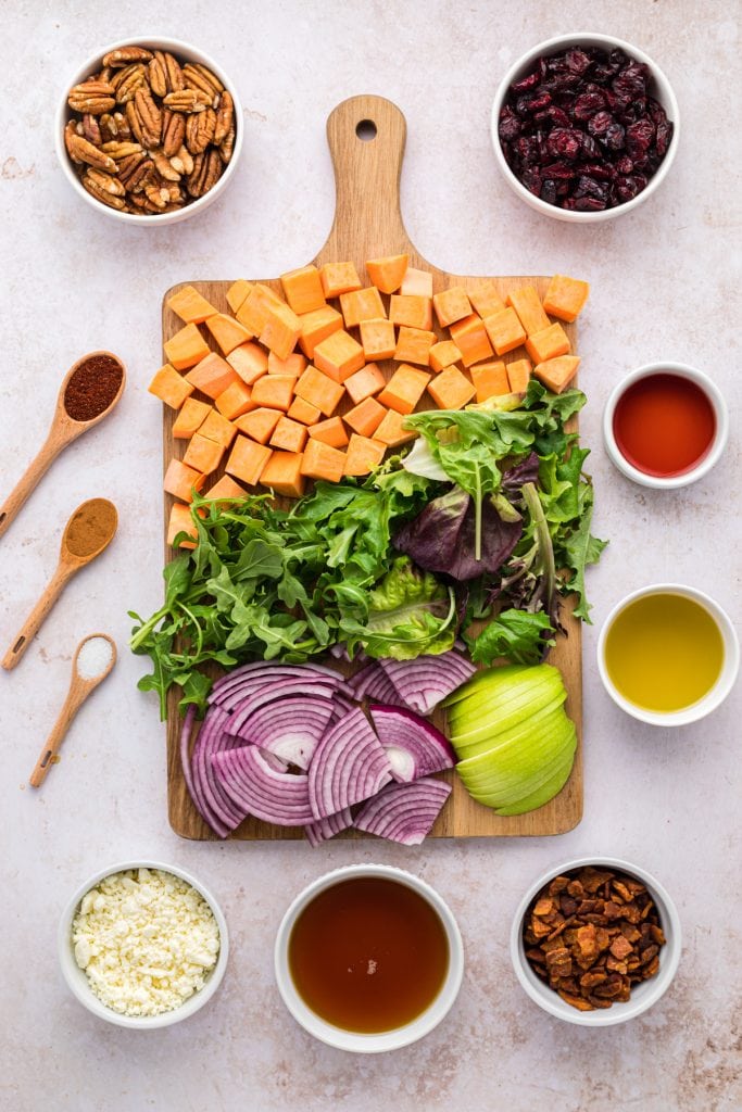 Picture of ingredients used with sweet potatoes, pecans, crumbled bacon, sliced apples, Craisins, mixed greens, feta cheese, sliced red onions, seasonings, and vinaigrette ingredients.