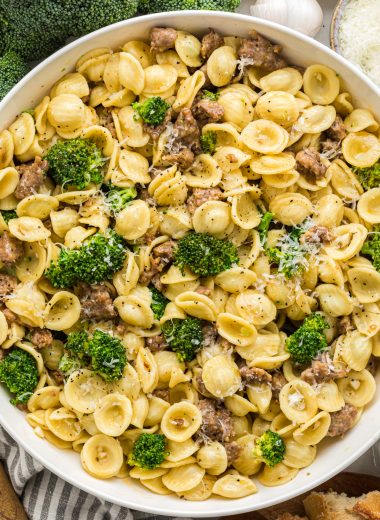 This easy Italian Sausage and Broccoli Pasta is a delicious weeknight meal on the table in 30 minutes. The sausage and broccoli cook in a flavorful light sauce seasoned with garlic, oregano, olive oil, chicken broth and cream. The pasta comes together with the addition of Parmesan cheese. Here the pasta is presented in a large serving bowl surrounded by ingredients used to make the dish including garlic, broccoli, salt and pepper, and Parmesan cheese accompanied with some fresh bread.