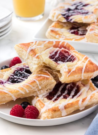 Delicate layers of puff pastry encase a cream cheese, berry, and jam filled danish. Each danish is glazed with a creamy almond kissed icing and contain a blueberry or raspberry filling. Here the pastries are stacked on a plate with a bite taken out of the top pastry.