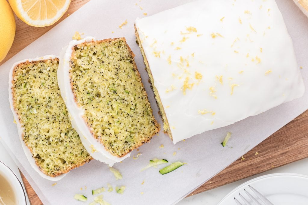 This moist, lemon zucchini poppy seed bread includes a lemon pudding mix, yogurt, shredded zucchini, lemon zest, and poppy seed in its ingredients. This delicious bread is covered in a creamy icing that has a touch of almond extract. Here is a picture of an overhead shot of the loaf with the first two pieces sliced.