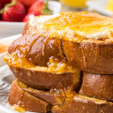This Orange French Toast with vanilla orange cream cheese and orange sauce is a beloved family favorite and the perfect holiday and weekend family breakfast. Here it is pictured stacked with 3 pieces of thickly sliced challah bread used for the french toast with the vanilla orange cream cheese on the top and orange sauce poured over the french toast and down the side.
