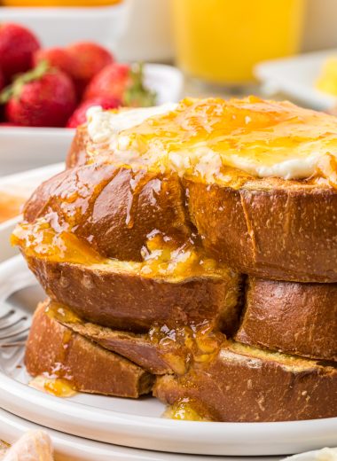This Orange French Toast with vanilla orange cream cheese and orange sauce is a beloved family favorite and the perfect holiday and weekend family breakfast. Here it is pictured stacked with 3 pieces of thickly sliced challah bread used for the french toast with the vanilla orange cream cheese on the top and orange sauce poured over the french toast and down the side.