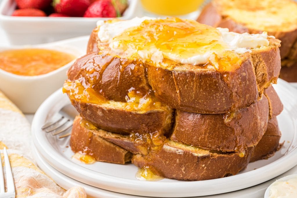 This Orange French Toast with vanilla orange cream cheese and orange sauce is a beloved family favorite and the perfect holiday and weekend family breakfast. Here it is pictured stacked with 3 pieces of thickly sliced challah bread used for the french toast with the vanilla orange cream cheese on the top and orange sauce poured over the french toast and down the side.