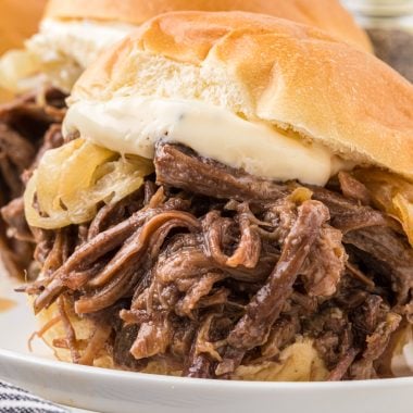 This recipes combines tender, flavorful shredded beef cooked in a slow cooker with caramelized onion and a garlic mayo dressing. Served in slider buns, they are the perfect family meal, appetizer for company, or a savory addition to your favorite sport watching party. In this image, two sliders are served on a plate with fries and a side of lemonade.