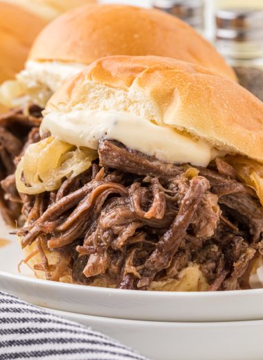 This recipes combines tender, flavorful shredded beef cooked in a slow cooker with caramelized onion and a garlic mayo dressing. Served in slider buns, they are the perfect family meal, appetizer for company, or a savory addition to your favorite sport watching party. In this image, two sliders are served on a plate with fries and a side of lemonade.