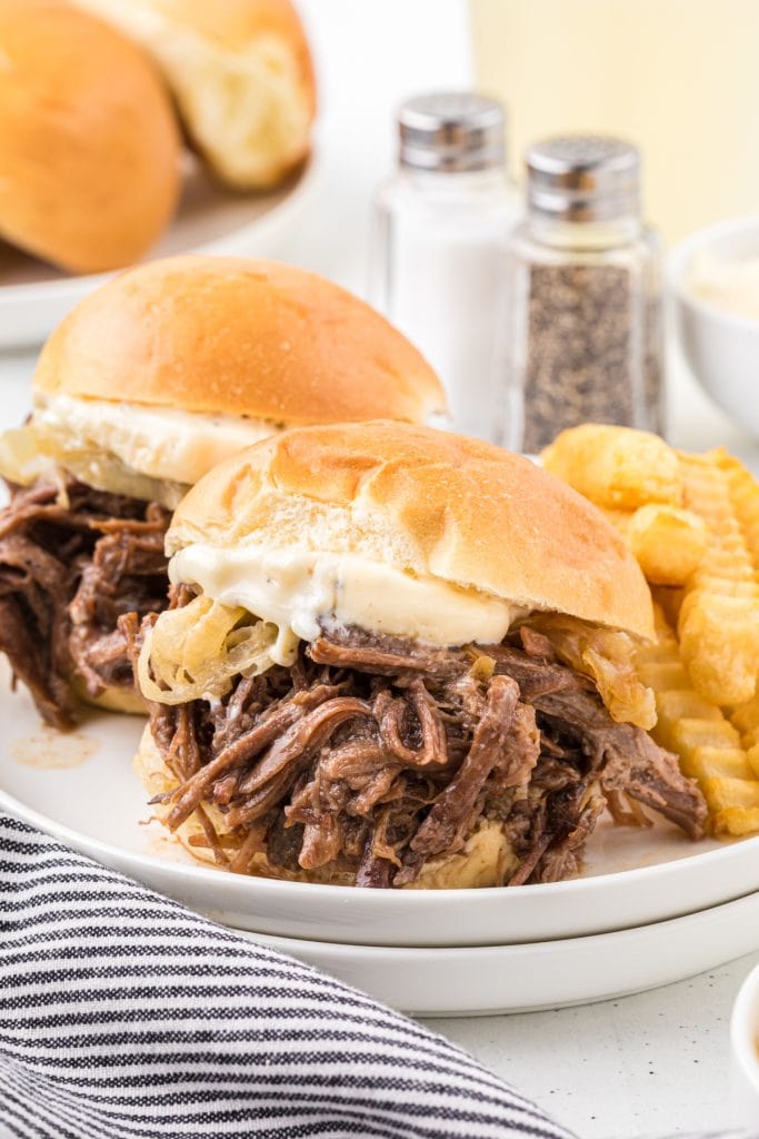 This recipes combines tender, flavorful shredded beef cooked in a slow cooker with caramelized onion and a garlic mayo dressing. Served in slider buns, they are the perfect family meal, appetizer for company, or a savory addition to your favorite sport watching party. In this image, two sliders are served on a plate with fries and a side of lemonade.