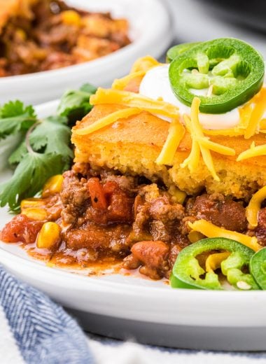 This Chili and Cornbread Casserole made with homemade chili seasoned with ground beef, chili beans, green chiles, broth, tomatoes, seasonings, and corn, and topped with a cornbread mix is an easy and delicious weeknight meal. We love the cornbread chili ratio and the slightly sweet cornbread combined with the flavorful seasoned chili. This casserole is tasty, healthy, comfort food sure to please kids and adults alike, it is a tried and true family favorite! Here it is pictured served on a dinner plate and garnished with sour cream, shredded cheese, sliced jalapeño and cilantro.