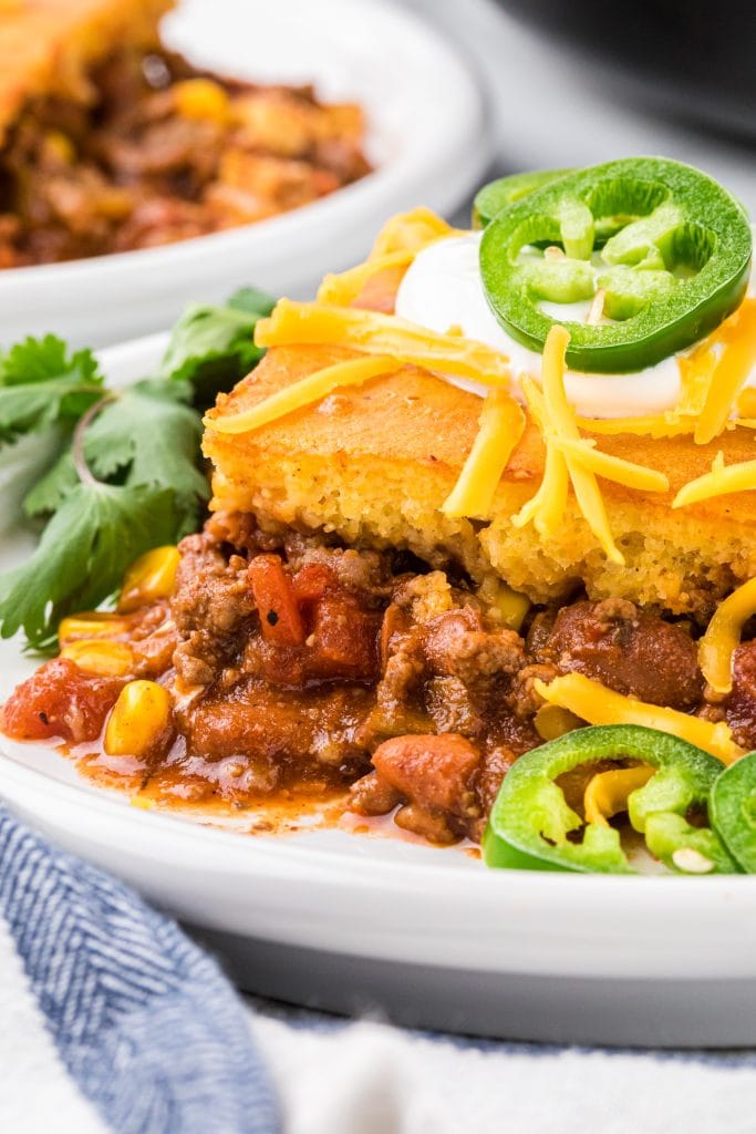 This Chili and Cornbread Casserole made with homemade chili seasoned with ground beef, chili beans, green chiles, broth, tomatoes, seasonings, and corn, and topped with a cornbread mix is an easy and delicious weeknight meal. We love the cornbread chili ratio and the slightly sweet cornbread combined with the flavorful seasoned chili. This casserole is tasty, healthy, comfort food sure to please kids and adults alike, it is a tried and true family favorite! Here it is pictured on a dinner plate garnished with sour cream, shredded cheese, jalapeño and cilantro.