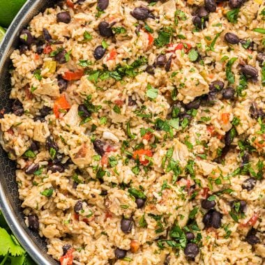 This flavorful Cilantro Lime Chicken with Rice has marinated cilantro lime chicken mixed with rice, onion, garlic, red pepper, black beans, tomatoes, green chiles, cilantro, cheese, and seasonings. Here the chicken and rice is pictured in the skillet it is made in with cilantro, lime, and garlic encircling the pan.