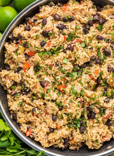 This flavorful Cilantro Lime Chicken with Rice has marinated cilantro lime chicken mixed with rice, onion, garlic, red pepper, black beans, tomatoes, green chiles, cilantro, cheese, and seasonings. Here the chicken and rice is pictured in the skillet it is made in with cilantro, lime, and garlic encircling the pan.