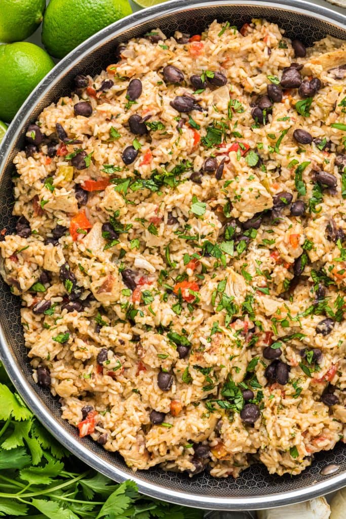 This flavorful Cilantro Lime Chicken with Rice has marinated cilantro lime chicken mixed with rice, onion, garlic, red pepper, black beans, tomatoes, green chiles, cilantro, cheese, and seasonings. Here the chicken and rice is pictured in the skillet it is made in with cilantro, lime, and garlic encircling the pan.
