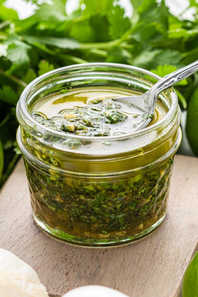 This easy Cilantro Lime Marinade for Chicken gives chicken incredible flavor in just 30 minutes. With just a few ingredients including cilantro, lime juice and zest, garlic, olive oil and seasonings, the chicken when cooked becomes tender, juicy, and flavorful, ready to be eaten alone or in a variety of dishes. Here the marinade is pictured in a small mason jar resting on a small cutting board surrounded by cilantro, garlic, and lime, with a spoon in the jar ready to dish out some marinade.