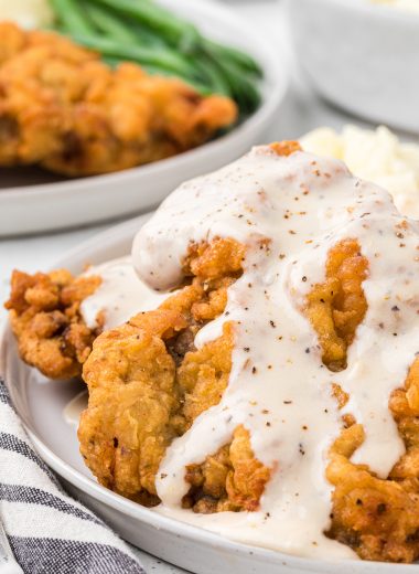 This Chicken Fried Steak with White Gravy is delicious Southern comfort food and easy to make at home. The seasoned, breaded cubed steak topped with a white country gravy is crispy and creamy perfection in the same bite. In this image, the prepared chicken fried steak is plated, topped with gravy and served with a side of mashed potatoes and green beans.