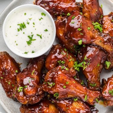 We love the ease and flavor of these Oven Barbecue Chicken Wings. The seasoned wings bake in the oven and then finish with an easy glaze of barbecue sauce with a touch of honey. They are the perfect crowd-pleasing wings for your next Game Day or gathering and are a family favorite. Here the wings are served on a plate with a side of ranch dressing.