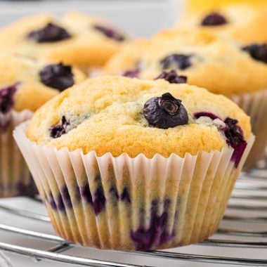 These Blueberry Buttermilk muffins are soft, sweet, and bursting with blueberries. The buttermilk in the muffins gives them an extra light and tender texture, and a touch of lemon zest and vanilla adds to the flavor with the blueberries. These muffins are a delicious treat for breakfast or brunch, or they make a favorite afternoon snack. Here the muffins are pictured cooking on a circular cooling rack with blueberries and orange juice in the background