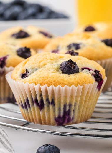These Blueberry Buttermilk muffins are soft, sweet, and bursting with blueberries. The buttermilk in the muffins gives them an extra light and tender texture, and a touch of lemon zest and vanilla adds to the flavor with the blueberries. These muffins are a delicious treat for breakfast or brunch, or they make a favorite afternoon snack. Here the muffins are pictured cooking on a circular cooling rack with blueberries and orange juice in the background