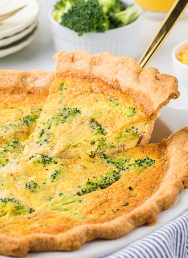 This Broccoli and Bacon Quiche is a breakfast favorite. The flaky crust, creamy custard, and bacon, broccoli, and cheddar cheese filling is rich, incredibly flavorful, and one of the best breakfast treats you can have. This is a perfect dish for a holiday brunch or a special weekend breakfast. In the picture, the quiche is pictured in the center with the first piece of the quiche being removed with a pie server and plates and juice in the background.