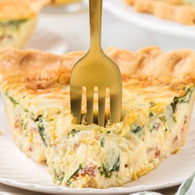 This Bacon and Spinach Quiche is a breakfast favorite. The flaky crust, creamy custard, and bacon, spinach, cheese, and onion filling is rich, incredibly flavorful, and one of the best breakfast treats you can have. This is a perfect dish for a holiday brunch or a special weekend breakfast. Here a slice of quiche is pictured plated, with a fork in the center of the first bite. The quiche and another plated piece are in the background.