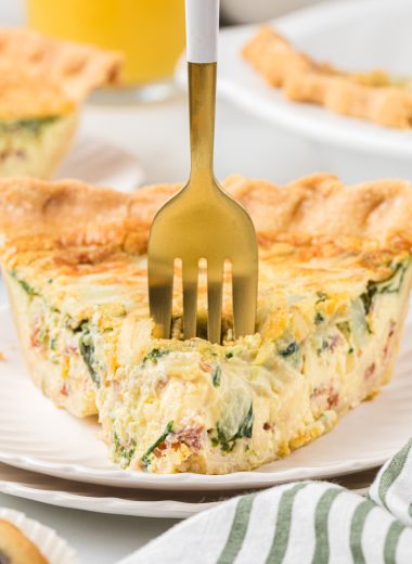 This Bacon and Spinach Quiche is a breakfast favorite. The flaky crust, creamy custard, and bacon, spinach, cheese, and onion filling is rich, incredibly flavorful, and one of the best breakfast treats you can have. This is a perfect dish for a holiday brunch or a special weekend breakfast. Here a slice of quiche is pictured plated, with a fork in the center of the first bite. The quiche and another plated piece are in the background.