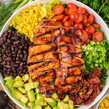 This Barbecue Ranch Chicken Salad with juicy barbecue chicken, smoky bacon, seasoned black beans, corn, tomatoes, creamy avocado, and onions is a delicious, filling main dish salad. Topped with homemade ranch dressing and a drizzle of barbecue sauce, it is a family favorite that kids and adults enjoy. Here the salad is arranged in a large bowl surrounded by additional ingredients from the salad, and ranch and barbecue dressing.