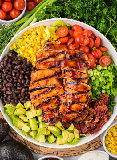 This Barbecue Ranch Chicken Salad with juicy barbecue chicken, smoky bacon, seasoned black beans, corn, tomatoes, creamy avocado, and onions is a delicious, filling main dish salad. Topped with homemade ranch dressing and a drizzle of barbecue sauce, it is a family favorite that kids and adults enjoy. Here the salad is arranged in a large bowl surrounded by additional ingredients from the salad, and ranch and barbecue dressing.