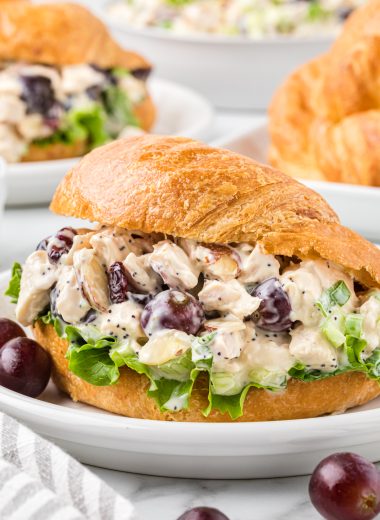 This is our favorite chicken salad. We love the chicken, celery, grapes, craisins, almonds, and green onions in the salad, and the dressing takes it over the top. The creamy mayonnaise poppy seed dressing has a little sugar, oil and vinegar, and Dijon mustard that really sets this chicken salad apart. Here the chicken salad is served on a plate in an open, halved croissant with additional croissants, sandwiches, and salad in the background.