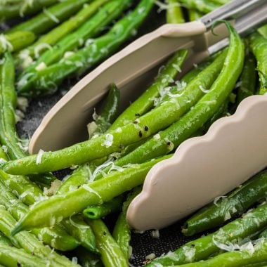 These Garlic Parmesan Green Beans are the perfect side dish for a weeknight meal or a special holiday dinner. Ready in minutes, these green beans are boiled to crisp-tender then sauteed with a delicious garlic butter seasoning and finished with a sprinkling of Parmesan. Here the prepared beans are pictured in a saute pan with tongs in the pan ready to pick up a serving of beans.