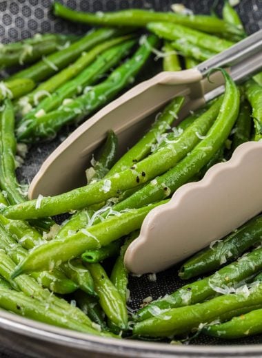These Garlic Parmesan Green Beans are the perfect side dish for a weeknight meal or a special holiday dinner. Ready in minutes, these green beans are boiled to crisp-tender then sauteed with a delicious garlic butter seasoning and finished with a sprinkling of Parmesan. Here the prepared beans are pictured in a saute pan with tongs in the pan ready to pick up a serving of beans.