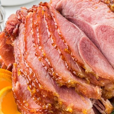 This Marmalade Glazed Ham is the perfect finishing touch for any holiday ham. The slightly sweet, citrusy, buttery glaze combined with the salty, smoky ham takes the ham to a whole new level. Ready in minutes, the glaze ingredients are whisked together and brushed on the ham during the last 20 minutes of baking. Here the ham is placed on a platter with orange and rosemary garnishes, with slices fanning out in front of the ham. Mashed potatoes and green beans are in the background.