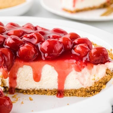 This Cherry and Cream Cheese Pie has a graham cracker crust, a no-bake cream cheese filling, and a sweet cherry pie topping. It is an easy dessert, perfect for the summer and winter, and is a favorite at family gatherings. In the image, the pie is cut into with the cut side facing the camera so the viewer sees the inside filling and crust with the cherry topping slightly spilling over the edge.