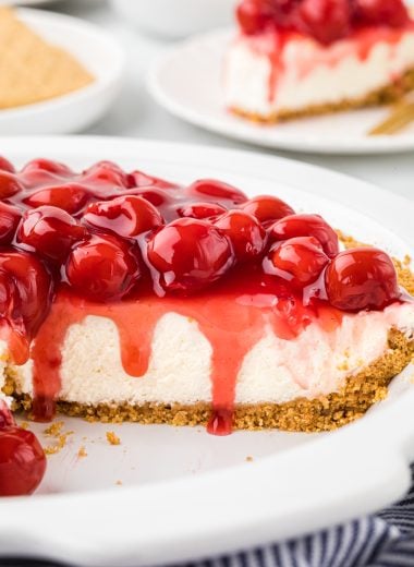 This Cherry and Cream Cheese Pie has a graham cracker crust, a no-bake cream cheese filling, and a sweet cherry pie topping. It is an easy dessert, perfect for the summer and winter, and is a favorite at family gatherings. In the image, the pie is cut into with the cut side facing the camera so the viewer sees the inside filling and crust with the cherry topping slightly spilling over the edge.