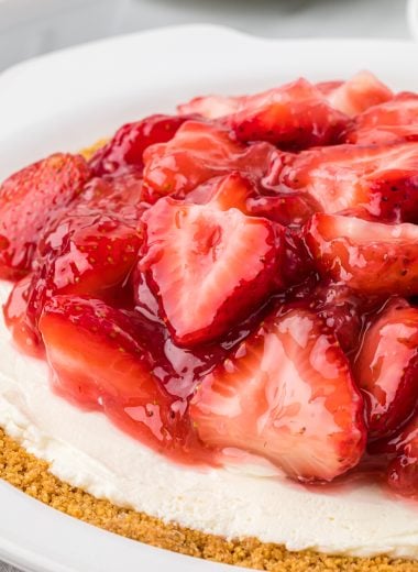 This Cream Cheese and Strawberry Pie has a graham cracker crust, a no-bake cream cheese filling, and a sweet homemade strawberry pie topping. It is an easy dessert, perfect for the summer holidays, and is a favorite at family gatherings. Here the whole pie is at the center of the image, prepared to serve, with the the focal point on the strawberry filling.