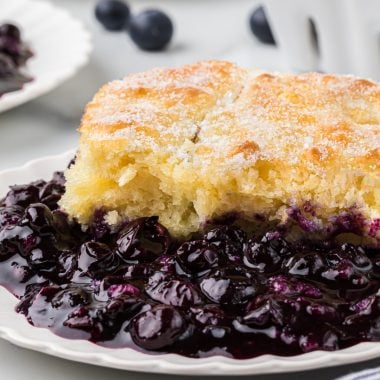 This Old-Fashioned Blueberry Cobbler has a tender buttermilk cake-like topping with a delicious, thick, and rich blueberry filling. We love the sprinkle of sugar on the slightly crisp top of the crust. This comforting dessert is a classic summer favorite and is always accompanied at our house with a scoop of vanilla ice cream. Here the cobbler is plated as an individual serving in the main center part of the frame with blueberries and other plates of cobbler in the background.