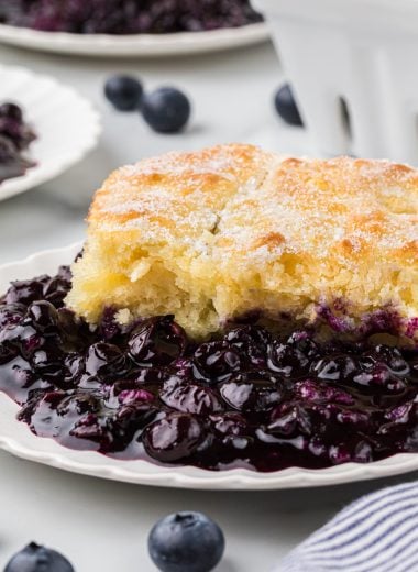 This Old-Fashioned Blueberry Cobbler has a tender buttermilk cake-like topping with a delicious, thick, and rich blueberry filling. We love the sprinkle of sugar on the slightly crisp top of the crust. This comforting dessert is a classic summer favorite and is always accompanied at our house with a scoop of vanilla ice cream. Here the cobbler is plated as an individual serving in the main center part of the frame with blueberries and other plates of cobbler in the background.