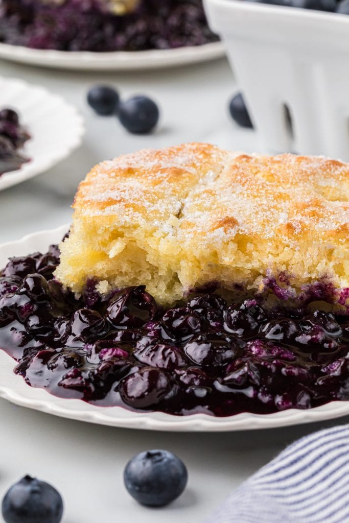 This Old-Fashioned Blueberry Cobbler has a tender buttermilk cake-like topping with a delicious, thick, and rich blueberry filling. We love the sprinkle of sugar on the slightly crisp top of the crust. This comforting dessert is a classic summer favorite and is always accompanied at our house with a scoop of vanilla ice cream. Here the cobbler is plated as an individual serving in the main center part of the frame with blueberries and other plates of cobbler in the background.