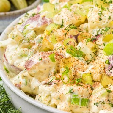 This Potato Salad with Dill Pickles is a favorite side dish for summer barbecuing. The salad has a creamy mayo based dressing mixed with red potatoes, celery, green onions, dill pickles, and hard-boiled eggs. It gets a flavor boost with a little Greek seasoning in the dressing and a dill and paprika garnish. Here the potato salad is featured in a serving bowl with dill, pickles and green onions on the side and background, all of which are ingredients in the salad or as a garnish.