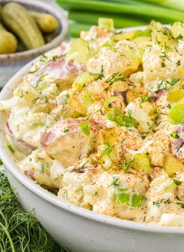 This Potato Salad with Dill Pickles is a favorite side dish for summer barbecuing. The salad has a creamy mayo based dressing mixed with red potatoes, celery, green onions, dill pickles, and hard-boiled eggs. It gets a flavor boost with a little Greek seasoning in the dressing and a dill and paprika garnish. Here the potato salad is featured in a serving bowl with dill, pickles and green onions on the side and background, all of which are ingredients in the salad or as a garnish.