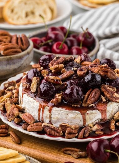 This delicious Baked Brie with Pecans and Cherry Sauce has a brown sugar coating on the brie, roasted pecans, and a sweet cherry vanilla balsamic sauce that perfectly complements the brie. We use frozen cherries to make it a year-round treat. It is one of my favorite appetizers; we love serving it with baguette slices or crackers. It is perfect served on its own or as part of a larger charcuterie board; it is both beautiful and delicious! Here the brie is plated with the cherry sauce and pecans on top of the Brie wheel and on the sides of the plate. The background pictures pecans, crackers, cherries, and sliced baguettes.