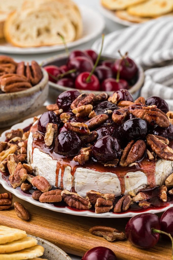 This delicious Baked Brie with Pecans and Cherry Sauce has a brown sugar coating on the brie, roasted pecans, and a sweet cherry vanilla balsamic sauce that perfectly complements the brie. We use frozen cherries to make it a year-round treat. It is one of my favorite appetizers; we love serving it with baguette slices or crackers. It is perfect served on its own or as part of a larger charcuterie board; it is both beautiful and delicious! Here the brie is plated with the cherry sauce and pecans on top of the Brie wheel and on the sides of the plate. The background pictures pecans, crackers, cherries, and sliced baguettes.