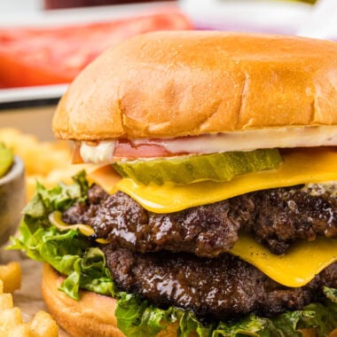 These smashburgers are our family's favorite burger. They cook in 4 minutes, have an amazingly juicy patty that locks in the flavor when smashed, a flavorful crisp crust, and a fantastic sauce that tastes so good with the burger. If you haven't tried making smashburgers before, these are a must try for your next barbecue! Here the smashburger is pictured centered in the image, assembled, and placed on a cutting board with fries in the sides and foreground and burger toppings and a drink in the background.
