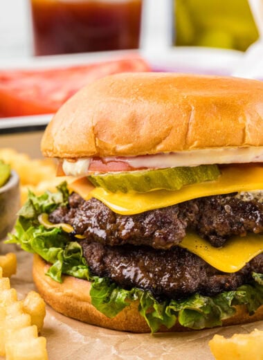 These smashburgers are our family's favorite burger. They cook in 4 minutes, have an amazingly juicy patty that locks in the flavor when smashed, a flavorful crisp crust, and a fantastic sauce that tastes so good with the burger. If you haven't tried making smashburgers before, these are a must try for your next barbecue! Here the smashburger is pictured centered in the image, assembled, and placed on a cutting board with fries in the sides and foreground and burger toppings and a drink in the background.