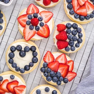 These Fruit Pizza Cookies with Berries have a sugar cookie base, cream cheese frosting, and an assortment of berries as a decorative topping. Decorating the cookies is fun for kids and adults alike, it's a great group or family activity, and a perfect 4th of July or summer treat. Here the cookies are displayed on a cooling rack, frosted and decorated with different patterns of berries with a linen and container of berries on the sides of the cooling rack.