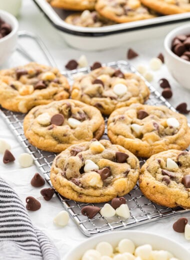 These Chocolate Chip and White Chocolate Chip Cookies are filled with three kinds of chocolate chips- semi-sweet, milk, and white. Golden and crisp on the outside with a soft and fudgy center, they have layers of chocolate flavor and are a huge family favorite. Here the cookies are centered in the image on a small cooling rack with white chocolate chips and chocolate chips scattered around the cookies and a tray of more cookies in the background.