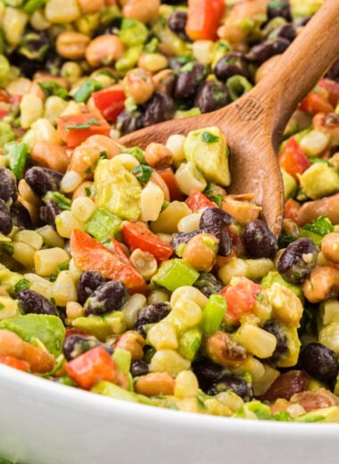 This Cowboy Caviar (aka Texas Caviar or Mississippi Caviar) is a classic summer side dish. Easy to prepare and full of flavor, it's a favorite choice for potlucks, barbecues, and get-togethers. Filled with black beans, black-eyed peas, tomatoes, corn, sweet peppers, avocado, green onion, and cilantro, and tossed with a lime and red wine vinegar dressing, we love serving this with tortilla chips or as a side with grilled chicken. Here the Cowboy Caviar is shown in a large bowl being scooped up with a wooden spoon in the center of the bowl.