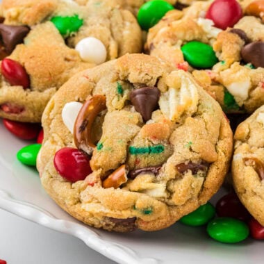 These Kitchen Sink Christmas Cookies are the ultimate salty-sweet combination cookie. Packed with red and green M&Ms, crushed pretzels, potato chips, chocolate chips, white chocolate chips, and red and green sprinkles, they are a festive, delicious cookie and my kids' absolute favorite. Here the cookies are plated in the center of the image with M&Ms and sprinkles in the foreground and background.