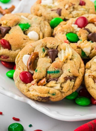 These Kitchen Sink Christmas Cookies are the ultimate salty-sweet combination cookie. Packed with red and green M&Ms, crushed pretzels, potato chips, chocolate chips, white chocolate chips, and red and green sprinkles, they are a festive, delicious cookie and my kids' absolute favorite. Here the cookies are plated in the center of the image with M&Ms and sprinkles in the foreground and background.