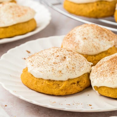 These Pumpkin Spice Cookies with Cream Cheese Frosting have all the flavors of Fall. They are soft, sweet, slightly spiced cookies with pumpkin pie spice, and the cream cheese frosting adds that special, sweet, creamy finish. In this image, there are three cookies on a plate in the center of the image with a cookie in the foreground, and another plate of cookies and a cooling rack with cookies in the background. A little pumpkin pie spice is sprinkled near the cookies.