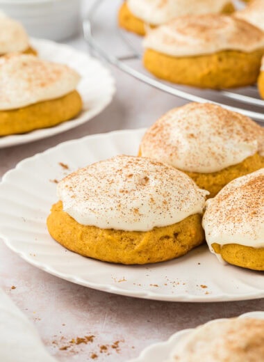 These Pumpkin Spice Cookies with Cream Cheese Frosting have all the flavors of Fall. They are soft, sweet, slightly spiced cookies with pumpkin pie spice, and the cream cheese frosting adds that special, sweet, creamy finish. In this image, there are three cookies on a plate in the center of the image with a cookie in the foreground, and another plate of cookies and a cooling rack with cookies in the background. A little pumpkin pie spice is sprinkled near the cookies.