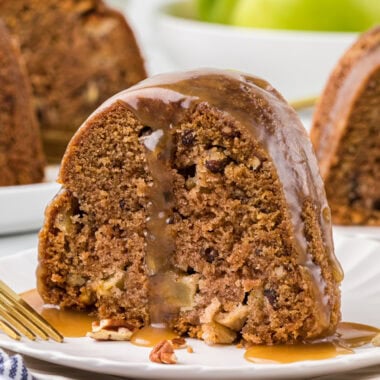 This Apple Caramel Bundt Cake is one of our favorite Fall desserts. The cinnamon-spiced cake is packed with tender pieces of apples and crisp pecans, and drizzled with a rich, homemade caramel sauce. In this image, a slice of the cake is plated in the center of the image with a fork resting on the plate and the caramel sauce drizzled over the top.