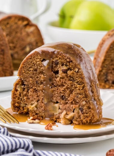This Apple Caramel Bundt Cake is one of our favorite Fall desserts. The cinnamon-spiced cake is packed with tender pieces of apples and crisp pecans, and drizzled with a rich, homemade caramel sauce. In this image, a slice of the cake is plated in the center of the image with a fork resting on the plate and the caramel sauce drizzled over the top.